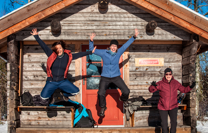 3 kvinnor hoppar framför en stuga i Gårdsjötorp